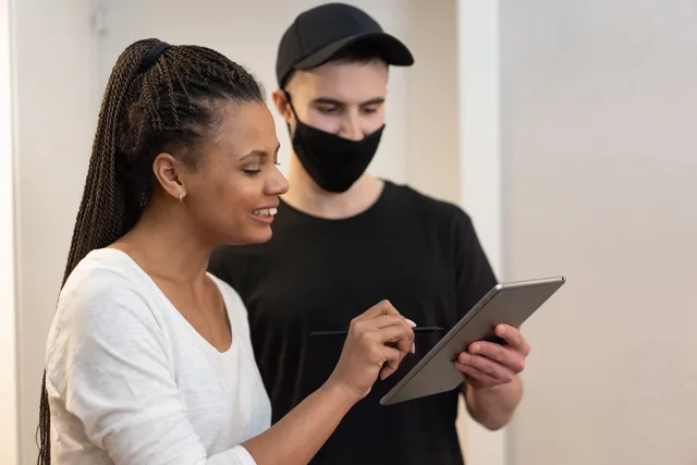 Woman signing for a delivery on a digital tablet held by a masked delivery man indoors. Signature électronique en Suisse : SCSE et valeur juridique des contrats